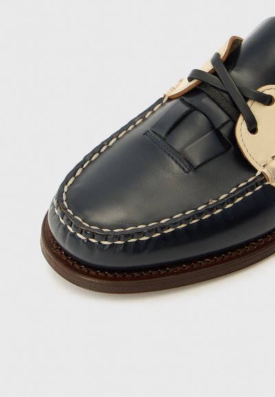 Scarpa da barca in pelle blu navy con accenti crema, cuciture bianche, suola piatta in gomma e design della punta arrotondata con chiusura a lacci.