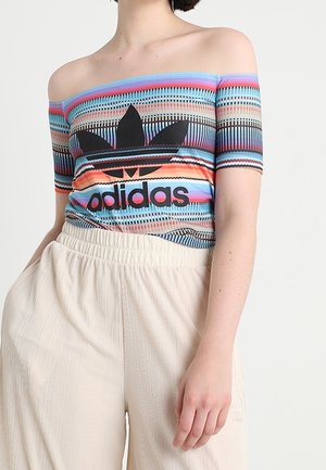 Nainen pukeutunut olkapäitä paljastavaan moniväriseen raidalliseen Adidas-paitaan, jossa on musta logo, yhdistettynä korkeavyötäröisiin kermaisiin leveälahkeisiin housuihin.