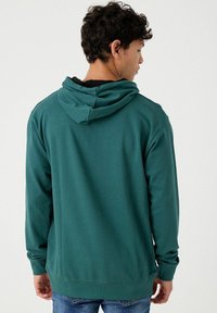Sudadera verde de tela suave con capucha ajustable, mangas largas y puños de canalé. Vista trasera que muestra un corte relajado y una textura suave.
