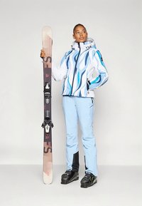 Skijas met een blauw golfpatroon, gecombineerd met lichtblauwe skibroek. De skiër houdt een ski met zwarte bindingen en bruine accenten vast. Zwarte skischoenen.