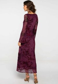Largo vestido burdeos con mangas largas transparentes, patrones florales en tonos más oscuros y una silueta fluida, combinado con sandalias de tacón con tiras.