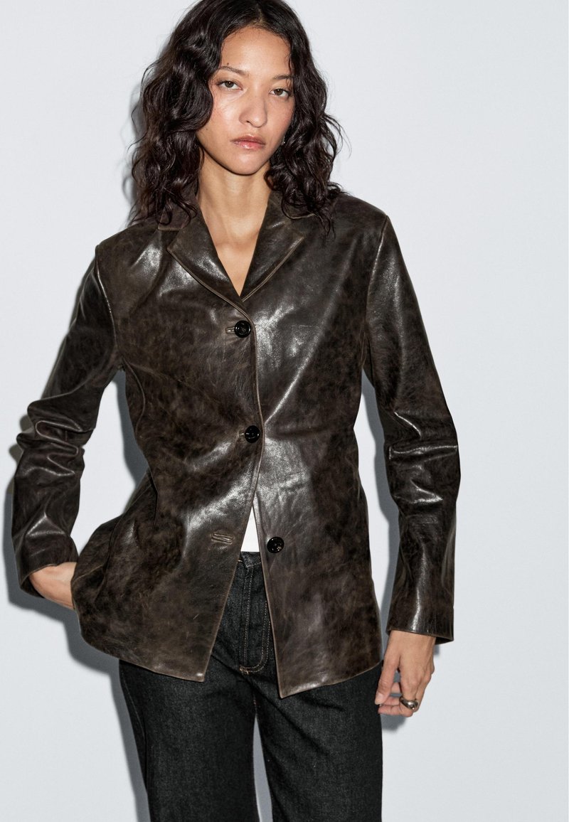 Vrouw met golvend donker haar, gekleed in een donkerbruine glanzende, knoopsluiting leren blazer en zwarte broek tegen een effen lichte achtergrond.