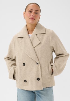 Femme portant un manteau en laine beige à double boutonnage avec les mains dans les poches et un jean bleu clair sur un fond blanc uni.