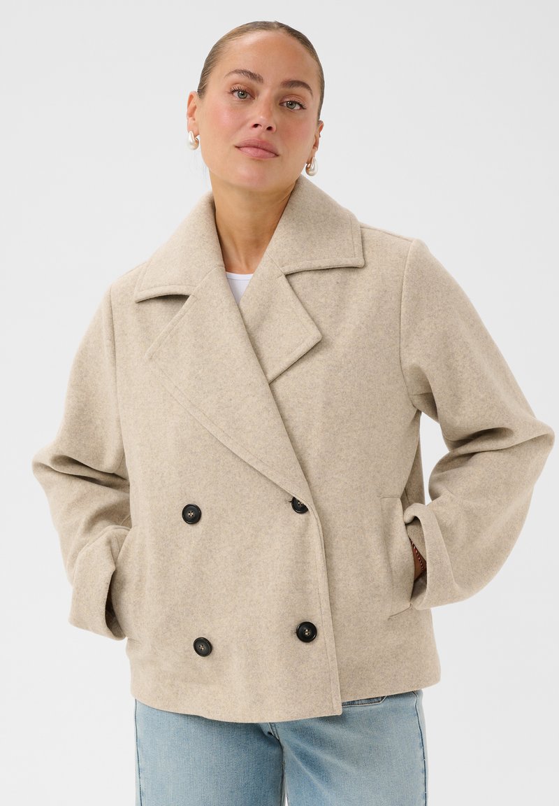 Femme portant un manteau en laine beige à double boutonnage avec les mains dans les poches et un jean bleu clair sur un fond blanc uni.