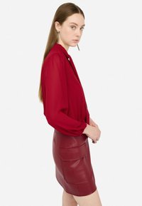 Blusa rossa con maniche lunghe a sbuffo abbinata a una gonna aderente in finta pelle rossa con tasche frontali. Texture liscia e design elegante.