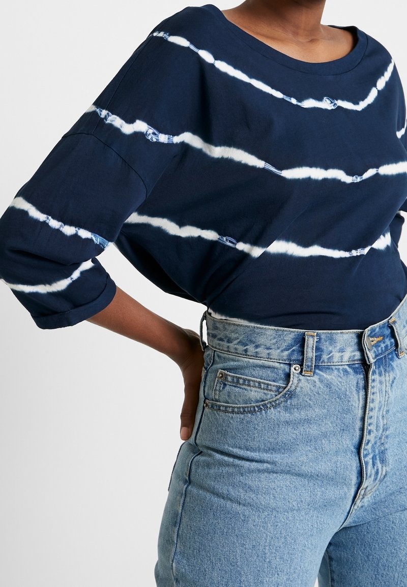 Blouse en coton bleu marine avec des rayures tie-dye blanches, encolure large et manches trois-quarts, associée à un jean taille haute bleu clair.