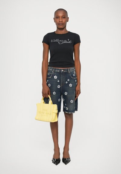 Schwarzes T-Shirt mit kurzen Ärmeln und weißem Logo, ausgefranste Denim-Shorts mit floralen Applikationen, gelbe Mini-Tote-Tasche und schwarze spitze Absätze.