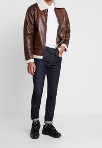 Bruine leren bomberjack met schapenvachtkraag, wit poloshirt, donkere denim jeans en zwarte sneakers. Het jack heeft een voorrits en manchetten.