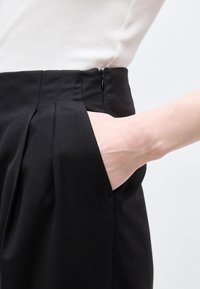 Χέρι που εισάγεται στην τσέπη μαύρου παντελονιού με ψηλή μέση, σε συνδυασμό με λευκό ριμπ τοπ.