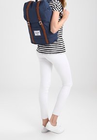 Granatowy plecak z brązowymi skórzanymi paskami i metalowymi akcesoriami. Prostokątny kształt z górnym klapą i przednią naszywką z logo. Nosi go osoba w paskowanej koszuli.