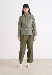 Olijfgroene gecapitonneerde jas met een ceintuur in de taille, vier voorzakken en een hoge kraag, gecombineerd met bijpassende losse olijfgroene broek en sneakers.