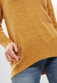 Pull en maille jaune moutarde avec poignets et ourlet côtelés, présentant une coupe ample et une texture douce. Associé à un jean en denim.
