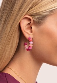 Pendientes de aro que presentan una mezcla de cuentas en tonos rosa y morado de varios tamaños, complementados con acentos dorados, exhibidos en la oreja de un modelo.