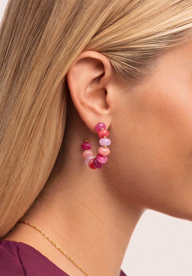 Pendientes de aro que presentan una mezcla de cuentas en tonos rosa y morado de varios tamaños, complementados con acentos dorados, exhibidos en la oreja de un modelo.