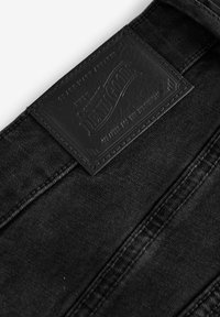 Zwarte denim jeans met een geprofileerde leren label waarop "DENIM GOODS" staat. Zichtbare stiksels langs de tailleband en naden.