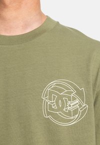 Olive green cotton t-shirt featuring a white graphic logo design on the left chest, round neckline, and short sleeves.