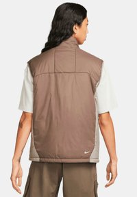 Brun vadderad väst med hög krage, som har grå sidodetaljer och en liten vit Nike-logotyp vid fållen. Strukturerad tyg, ärmlös design.