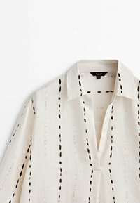 Camicia bianca in lino con colletto, caratterizzata da dettagli ricamati neri verticali e vestibilità rilassata. Maniche lunghe e design minimalista.