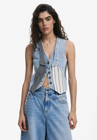 Chaleco de mezclilla con una mezcla de colores a rayas y sólidos, con escote en V, cierre de botones y un ajuste relajado. Unos pantalones vaqueros azul liso completan el look.