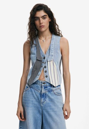 Gilet en denim avec un mélange de couleurs rayées et unies, présentant un col en V, une fermeture à boutons et une coupe décontractée. Un jean bleu uni vient compléter le look.
