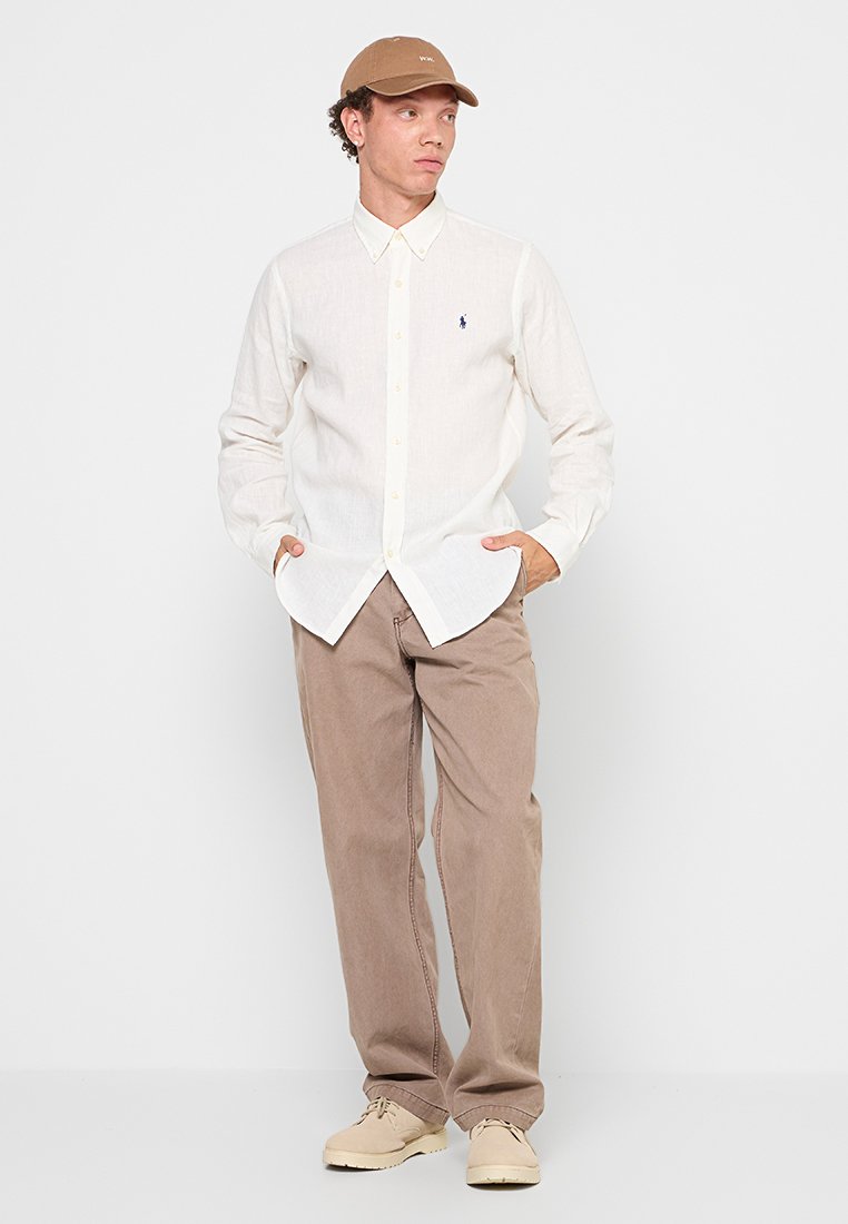 Chemise blanche à boutons avec col, pantalon beige et chaussures crème. La chemise présente un petit logo sur la poitrine gauche et des manches longues.