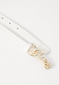 Ceinture en cuir blanc avec une boucle en métal doré portant le logo "Guess", dotée d'une texture lisse et de plusieurs trous pour régler la taille.