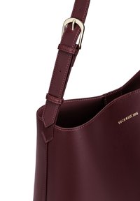 Burgundy leather tote bag with a smooth texture, adjustable strap, and gold-tone hardware. Features subtle branding on the side.