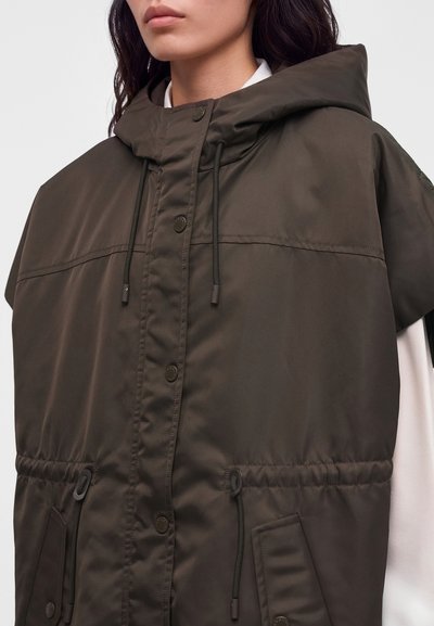 Femme portant une veste sans manches à capuche de couleur vert olive foncé avec des boutons sur le devant et des cordons à la capuche et à la taille.