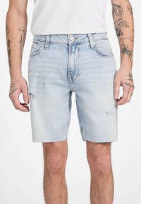 Shorts en denim bleu clair avec un design déchiré, pr�ésentant des ourlets effilochés et des détails de couture visibles. Portés avec un t-shirt blanc uni.