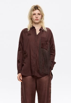 Brown shirt with floral embroidery, pocket detail, and fringe. The fabric appears smooth with a slightly shiny finish. Paired with satin-like brown pants.