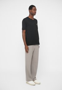 Μαύρο κοντομάνικο μπλουζάκι V με χρυσό λογότυπο στο αριστερό στήθος, συνδυασμένο με ανοιχτό γκρι πλισέ παντελόνι και λευκά αθλητικά παπούτσια.