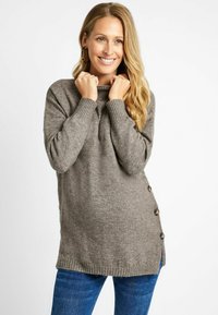 Pull tricot gris avec un col montant, des poignets côtelés et des boutons sur le côté. Texture douce, coupe légèrement ample et manches longues. Associé à un jean bleu.