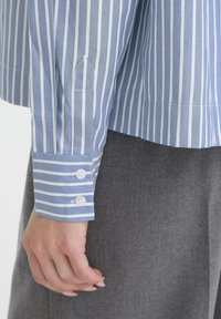 Hand neben grauen Hosen, trägt ein blau-weiß gestreiftes Langarmhemd mit geknöpften Manschetten.