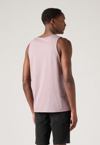 Svijetlo ružičasti tank top od pamuka, s opuštenim krojem i okruglim izrezom, uparen s crnim traper kratkim hlačama. Stražnji pogled prikazuje detalje ramena i ruku.