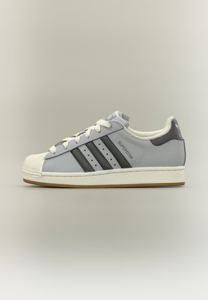 Sneaker Adidas Superstar basso grigio con strisce nere, suola crema, lacci bianchi e punta sagomata.