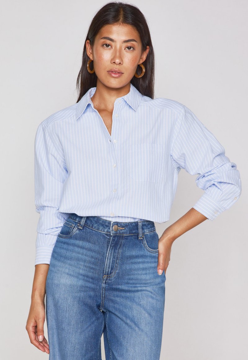 Camicia a righe blu chiaro con colletto, maniche lunghe arrotolate ai polsi e tasca applicata. Abbinata a jeans a vita alta.