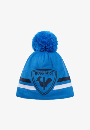 Gorro de punto azul con un gran pompón en la parte superior. Presenta un patrón de rayas en blanco y negro y un logotipo de un gallo dentro de un escudo.