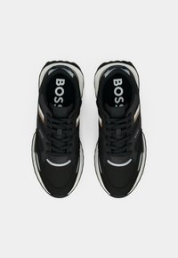 Baskets noires avec une tige en mesh et en synthétique, présentant des accents blancs et métalliques, une semelle extérieure blanche épaisse et un branding visible sur la languette.