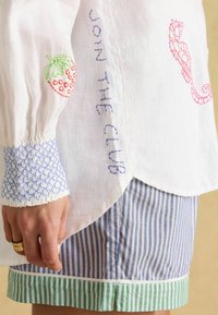 Hemd aus weißem Leinen mit buntem gesticktem Erdbeermotiv, Seepferdchen und dem Text "JOIN THE CLUB"; mit blauen gemusterten Manschetten und gestreiften Shorts mit grünem Saum.