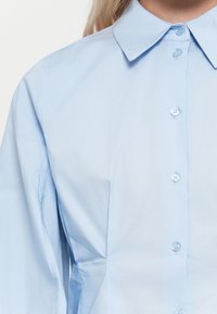 Camicia di cotone azzurra chiara con colletto, chiusura frontale con bottoni e maniche lunghe con dettagli di plissettatura discreti. Texture liscia.
