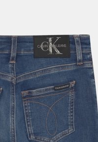 Vaqueros de mezclilla azul oscuro con una etiqueta de tela negra que presenta "Calvin Klein Jeans" y un logo metálico "CK". Costuras en contraste visibles.