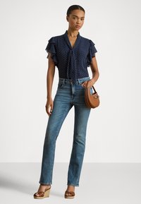 Nő áll, kék pöttyös blúzban fodros ujjakkal, világoskék farmerben, barna ék szandálban, kezében egy kis barna kézitáska.