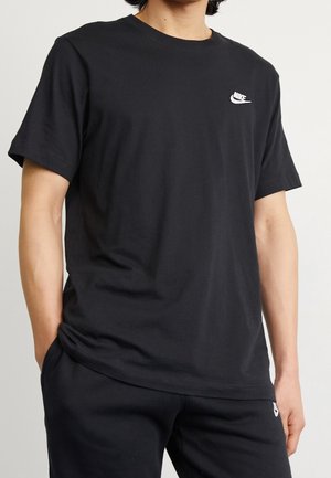 Person wearing a black Nike T-shirt with a white logo on the chest and matching black pants, standing with hands in pockets.