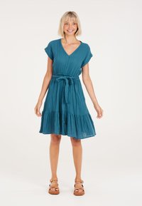 Teal katoenen jurk met korte opgerolde mouwen, V-hals, strikceintuur in de taille en een gelaagde, gerimpelde zoom. Zachte textuur. Sandalschoenen.