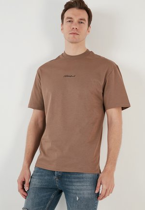 Brown cotton t-shirt with a crew neck, short sleeves, and black scripted logo on the chest, worn with blue distressed jeans.