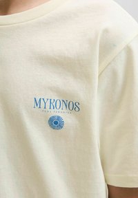 Camiseta de algodón color crema, con el texto impreso en azul "MYKONOS PURE PARADISE" y un gráfico circular en la parte izquierda del pecho.