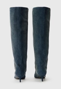 Botas de mezclilla por encima de la rodilla con un diseño ajustado, color azul oscuro, punta puntiaguda y tacón delgado y estable. Tela texturizada con costuras visibles.