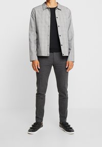 Šedá kostkovaná bunda na knoflíky přes černé tričko, doplněná šedými texturovanými kalhotami a černými teniskami s pásky.