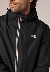 Man in een zwarte hooded jacket met witte rits en het "Deeluxe Original" logo op de borst, kijkend naar de zijkant tegen een effen achtergrond.