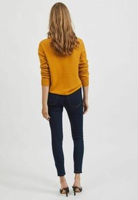 Maglione giallo a maglia con polsini a costine, abbinato a jeans skinny blu scuro. L'outfit è completato da sandali neri con tacco alto.